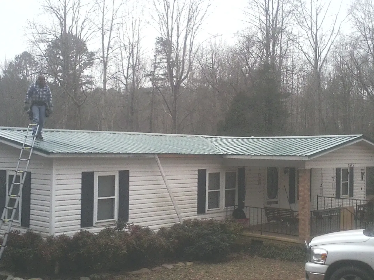 Worker on a ladder beside a new green metal roof on a ranch home in Church Point
