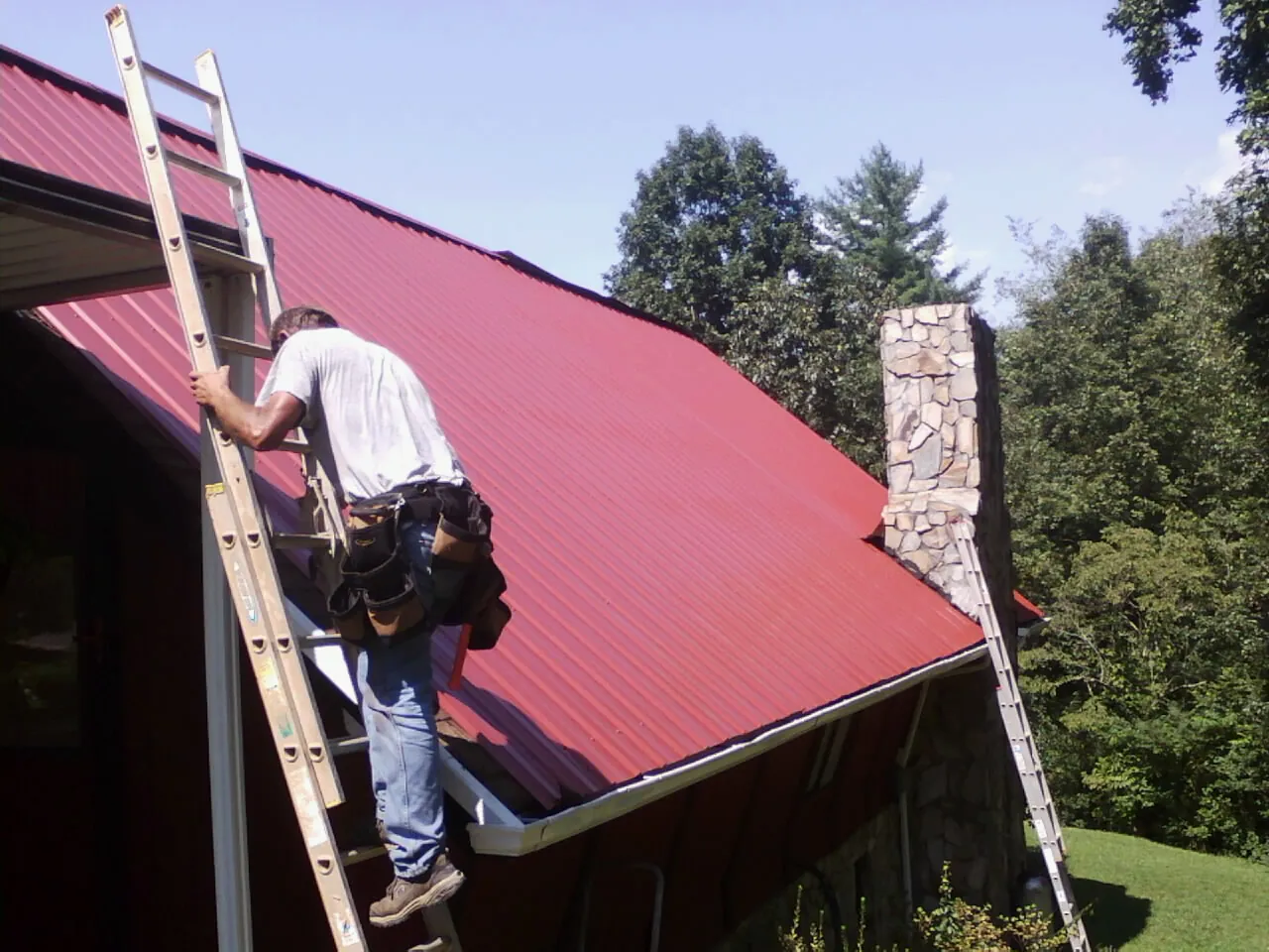 Roofer on a ladder beside a red metal roof and stone chimney — Soffit & Fascia Repair by Roofing Brothers in Church Point